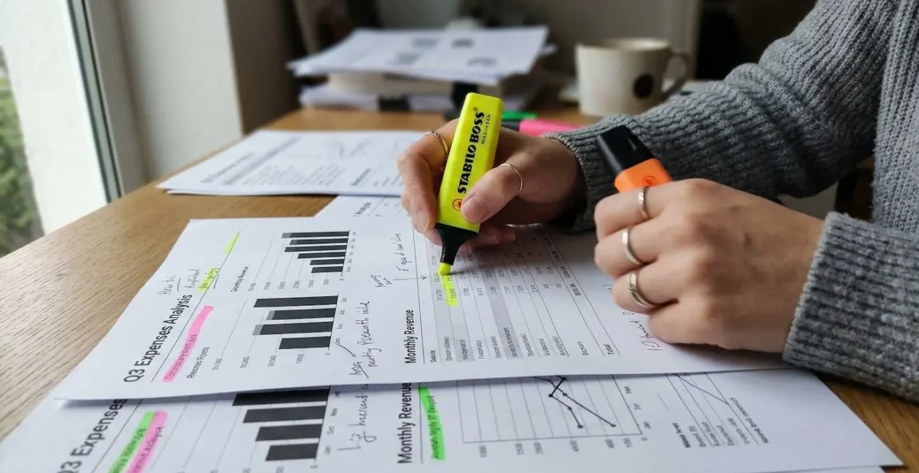 Des mains parcourent un dossier rempli de graphiques chiffrés avec des surligneurs colorés sur un bureau de travail.